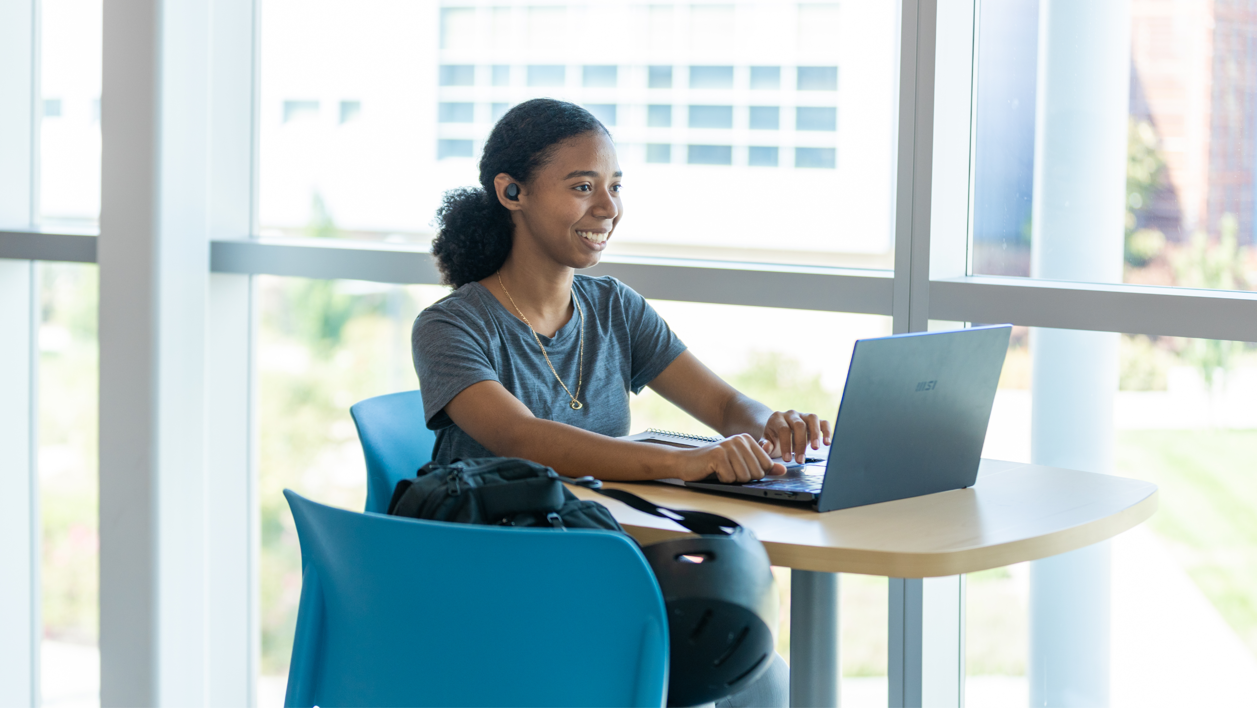 A female student sitting at a tall table near a bright window, smiling as she works on a laptop with earbuds in.