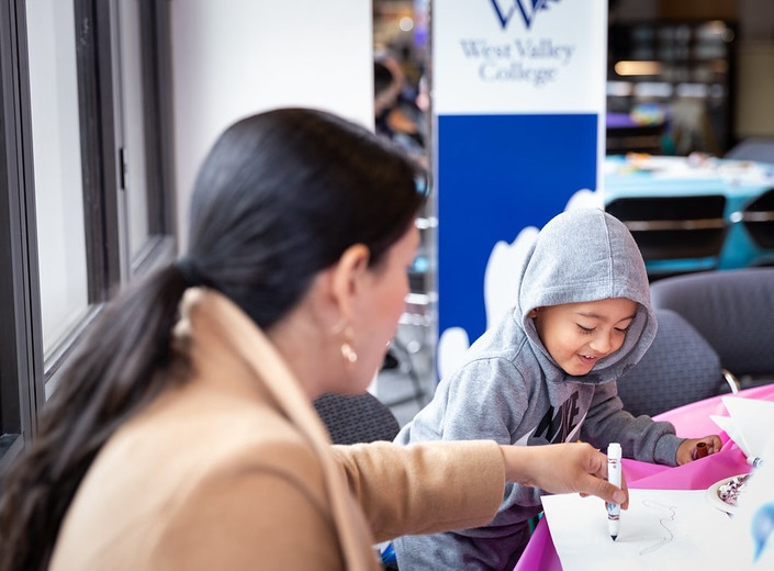 Latina with long black hair in ponytail faces away from camera and helps a young boy draw.