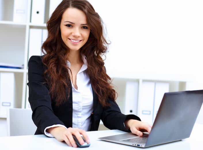 Young business woman in a blazer works at a laptop. She has long brown hair.