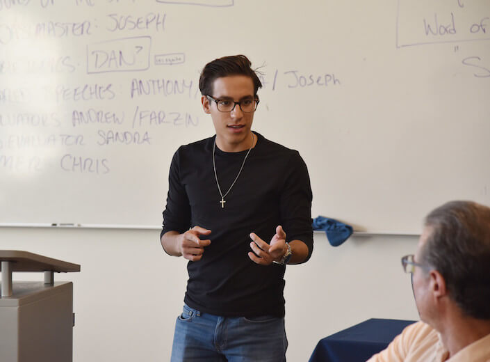 A young man delivers his speech in Toastmasters. He has olive skin, short black hair, and dark-rimmed glasses.