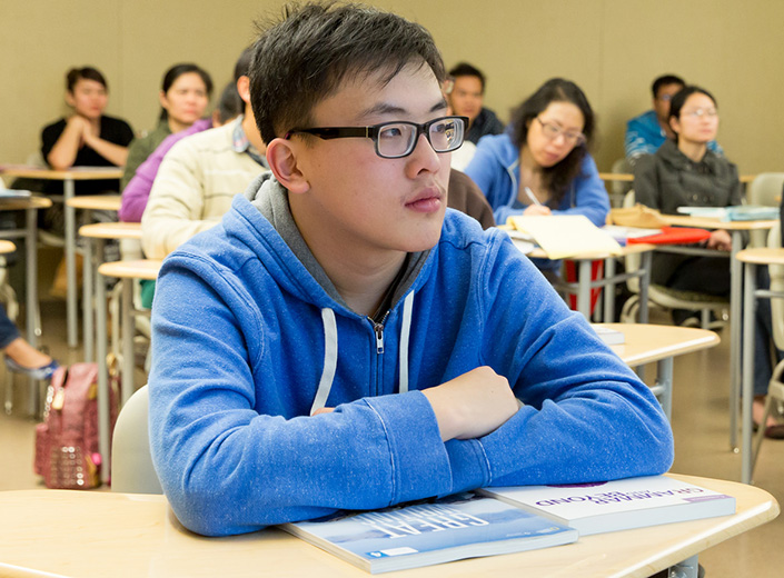 Young Asian man in blue hoodie and black frame glasses sits in college classroom.