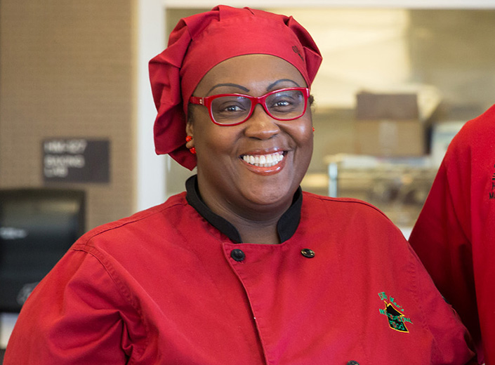 Female Hospitality Management student works in a food truck. She wears a chef's uniform and glasses. 