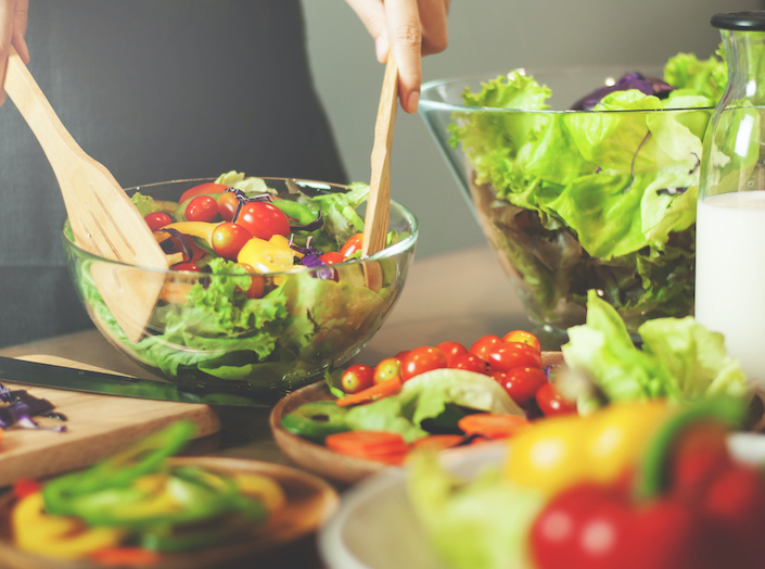 Fresh vegetables are laid out on a table to be prepared as a salad. A woman's hands hold salad tongs and she mixes the ingredients in a large bowl.