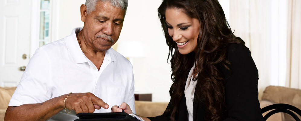 A woman with dark hair assists an elderly latino man with a process on a black tablet.