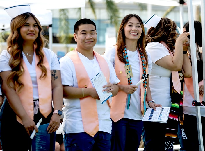 Three student nurses pose in their white scrubs with peach-colored sashes.