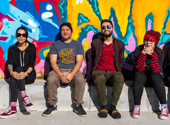 Mural project. Three students sit with instructor on curb in front of a bright mural. One female/slim/Latina in a black hoodie, one slim and white or Latino with green pants and red shirt, and the other female with bright red pink hair under a maroon beanie and striped red and black long-sleeved shirt. The instructor is in a t-shirt with a yellow/red logo (non discernible), a black beanie, and dark tan pants. He is Latino and in late twenties/thirties.