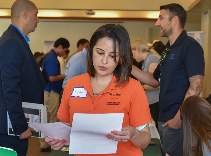 Career Center - a young woman with shoulder-length black hair and an orange shirt participates in a career fair. She is holding and looking at a few papers. There are people behind her engaged in conversations.