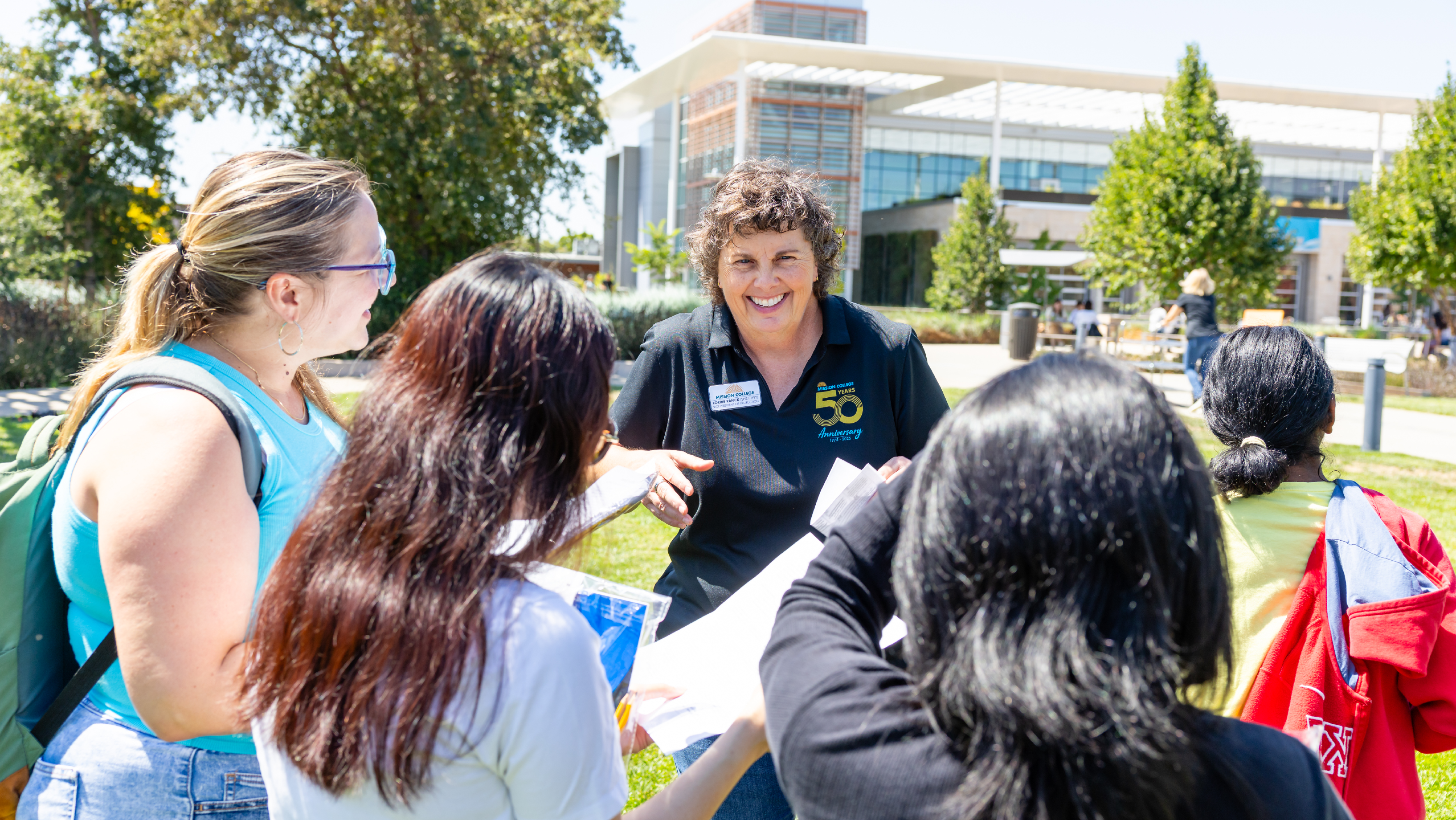 A smiling staff member wearing a black Mission College 50th Anniversary polo shirt handing out papers to a small group of students on a sunny campus lawn.