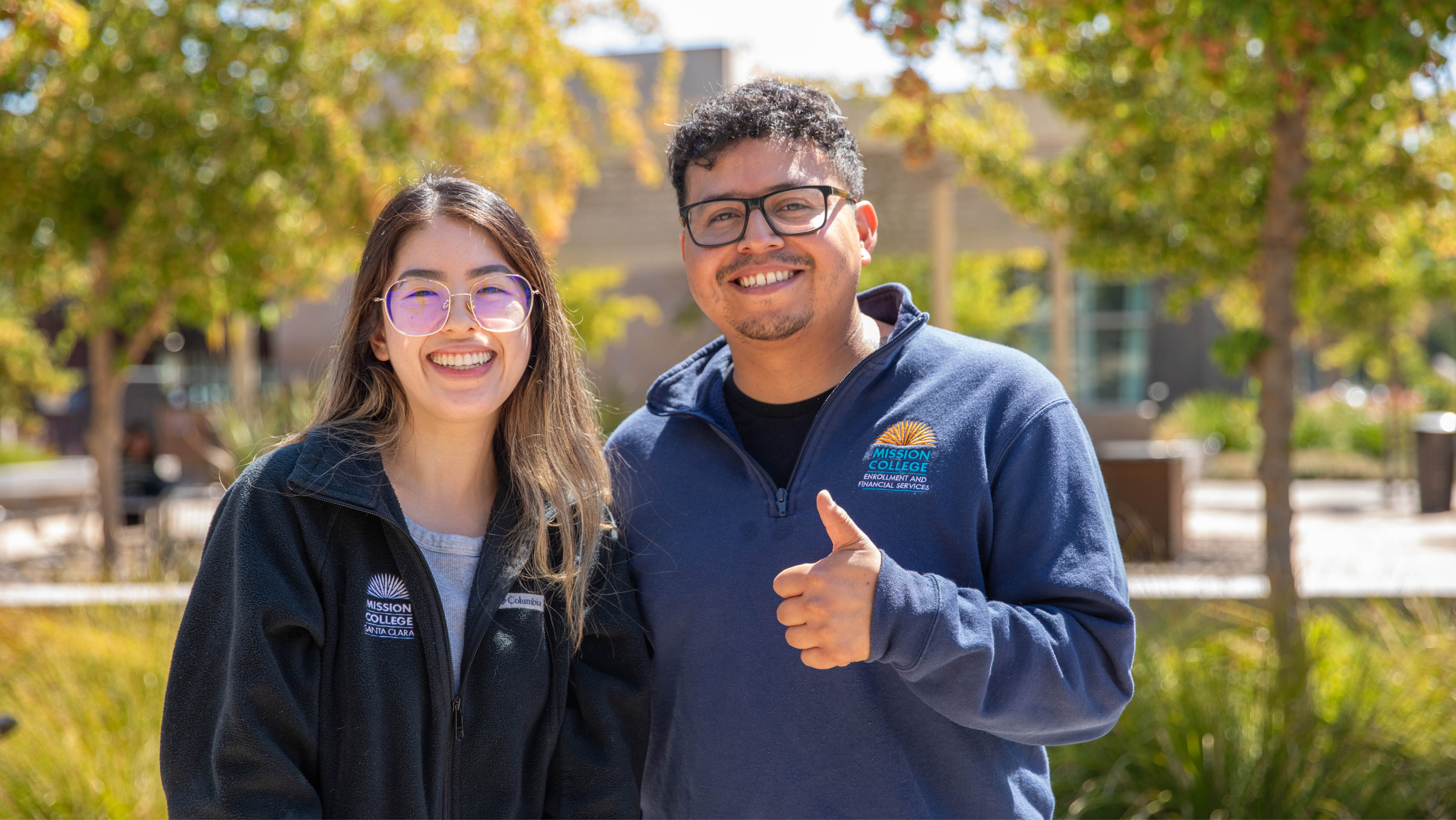 Two smiling students wearing Mission College jackets outdoors; the man on the right gives a thumbs-up to the camera.