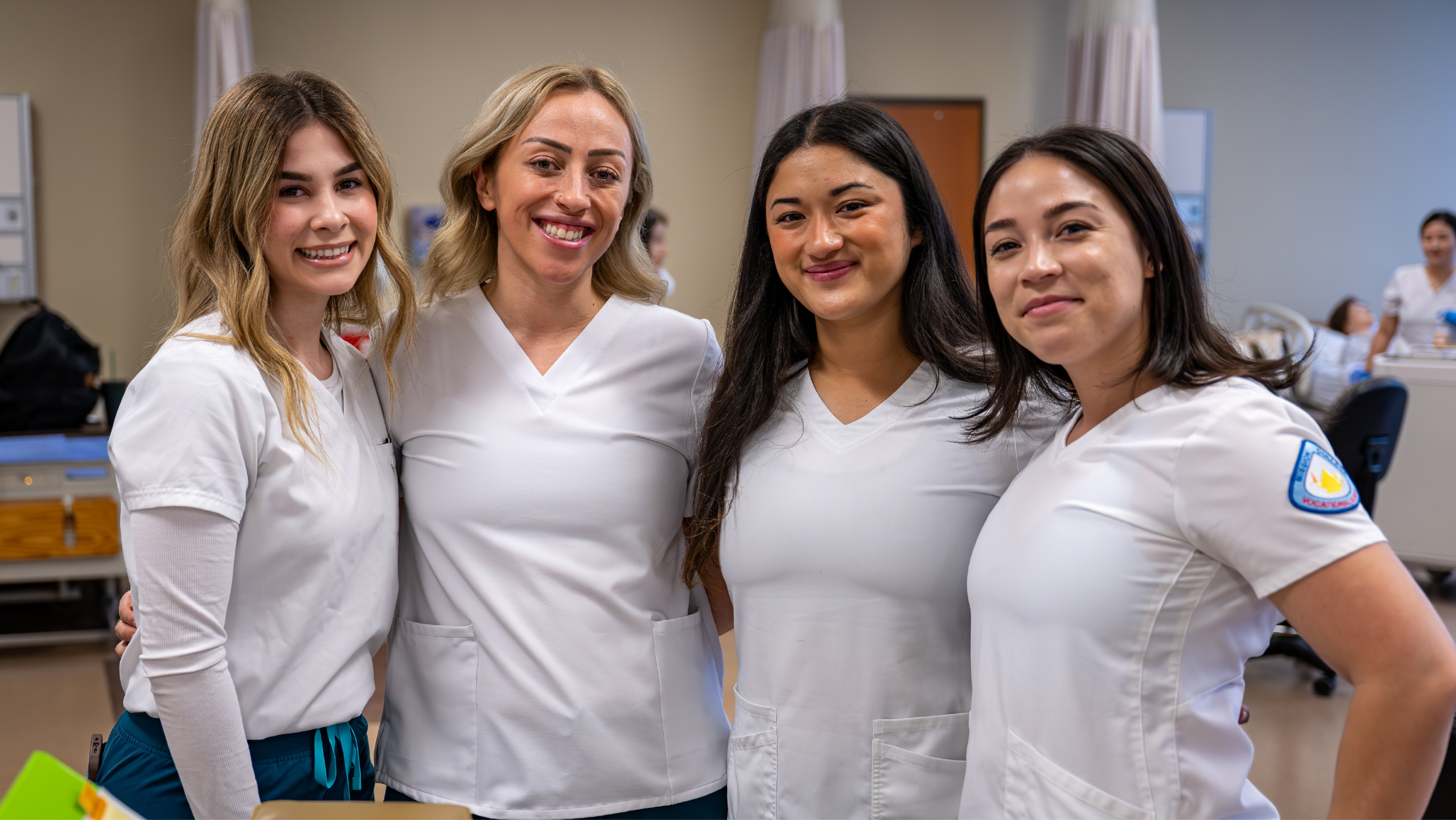 Four female nursing students smiling together indoors while wearing matching white scrubs inside a medical training facility.
