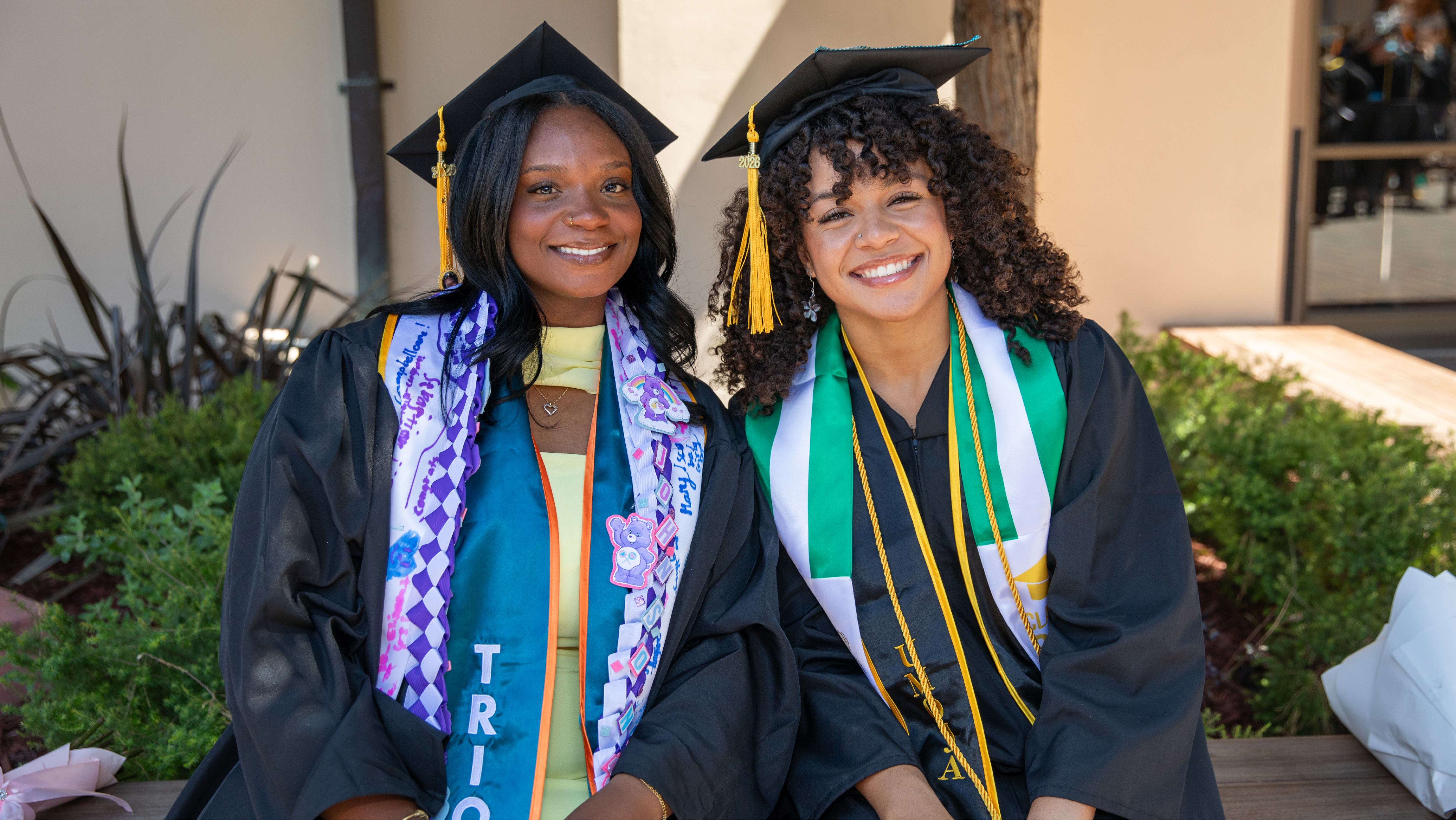 Two smiling female graduates sitting outdoors in black caps and gowns decorated with colorful graduation sashes and leis.