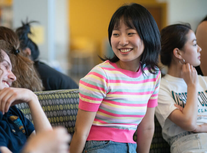 Young woman of Asian descent with a shoulder-length black bob smiles. She leans forward as she looks toward amother student off-camera and smiles. She is surrounded by other students, they sit in the lobby of the Student Engagement Center.