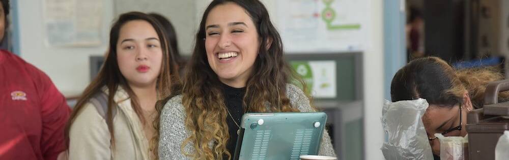 Girl with laptop in line for coffee at a school event. She has curly light brown hair, tan skin, and wears a sweater. She is laughing.