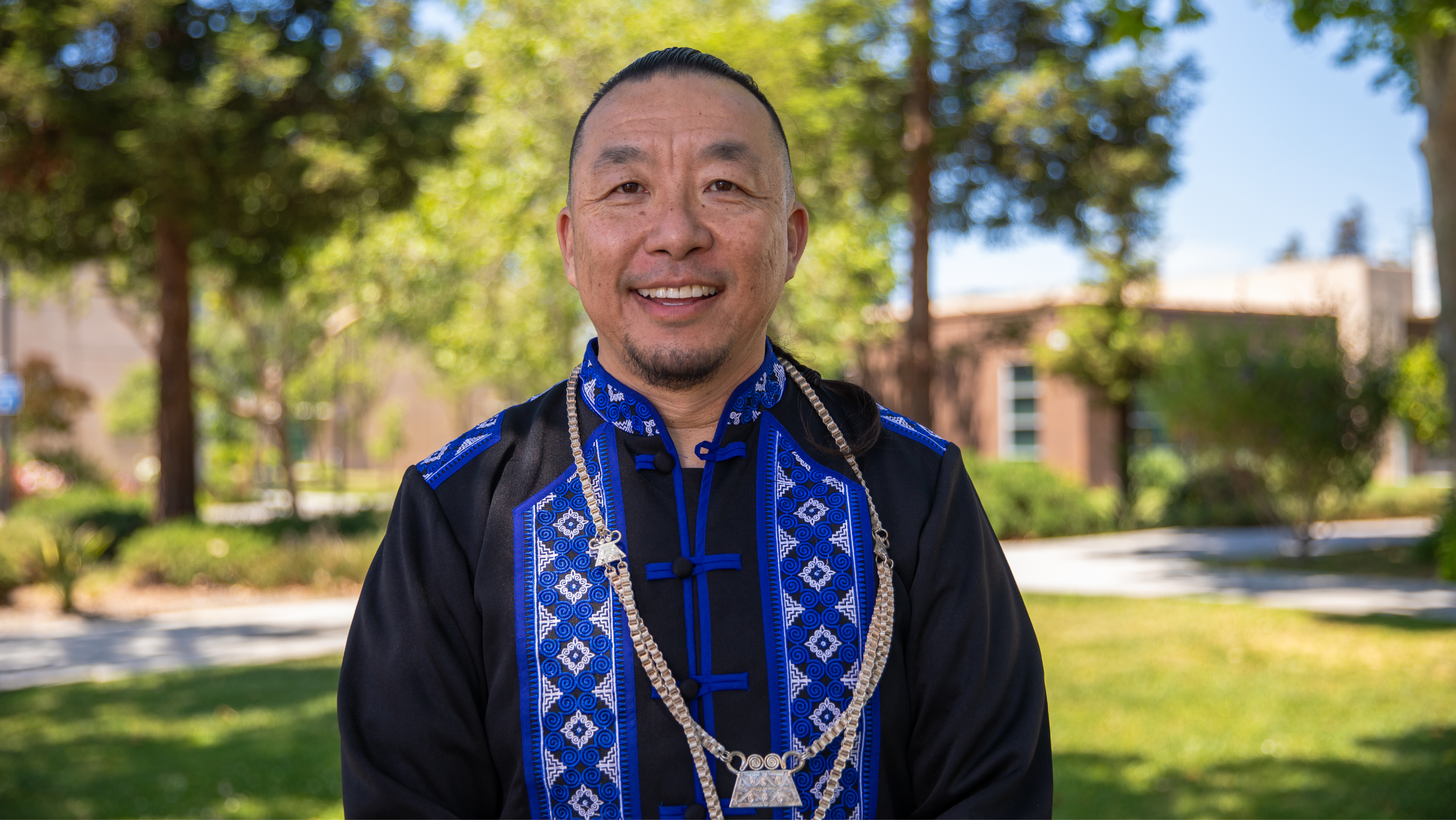 Close-up portrait of a smiling man outdoors wearing a traditional black and blue embroidered shirt paired with a silver lock necklace.