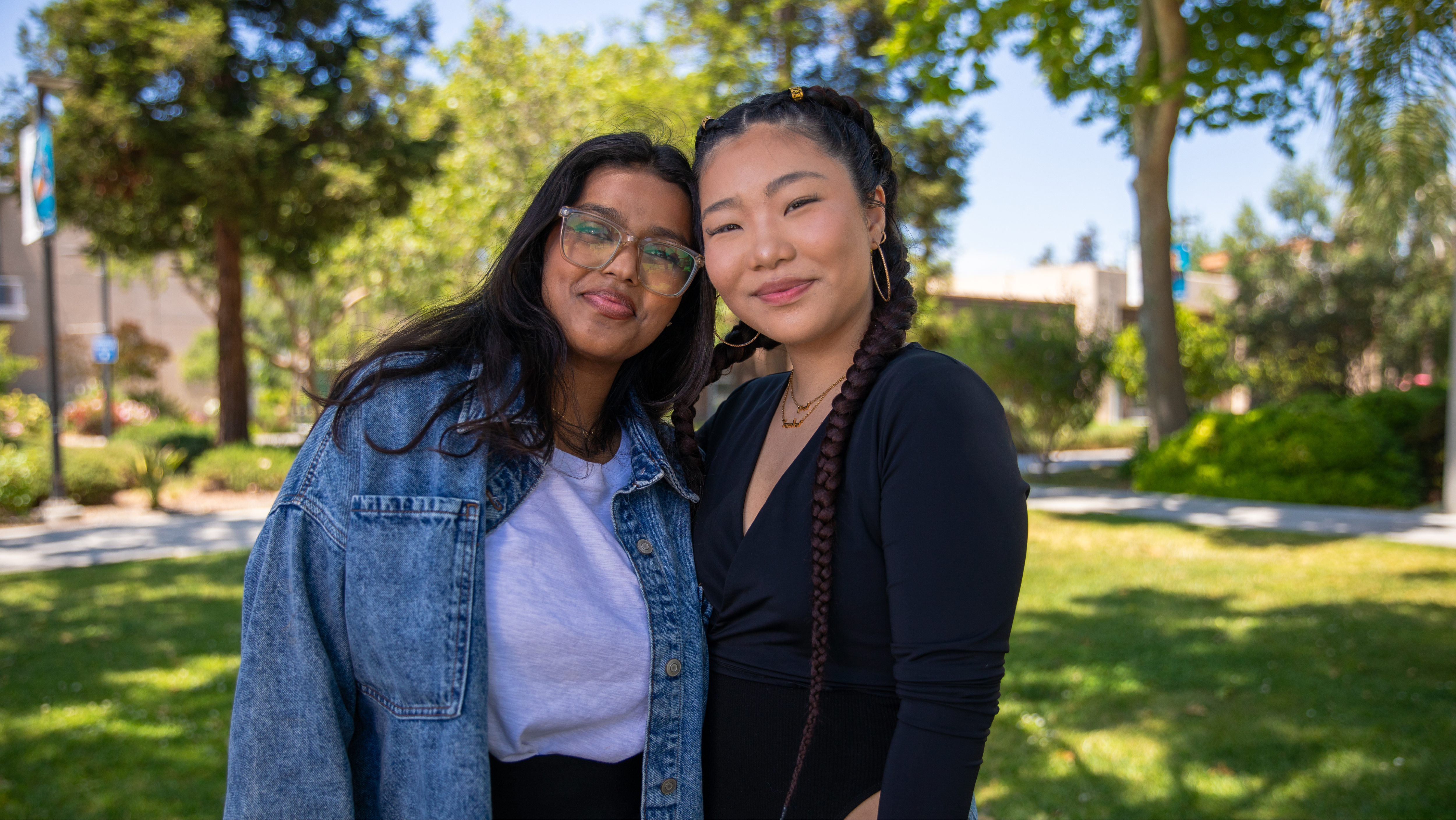 Two young women smiling together outdoors on a sunny campus lawn; one wears a denim jacket over a white top with glasses, while the other wears a black wrap top and hoop earrings with braided hair.