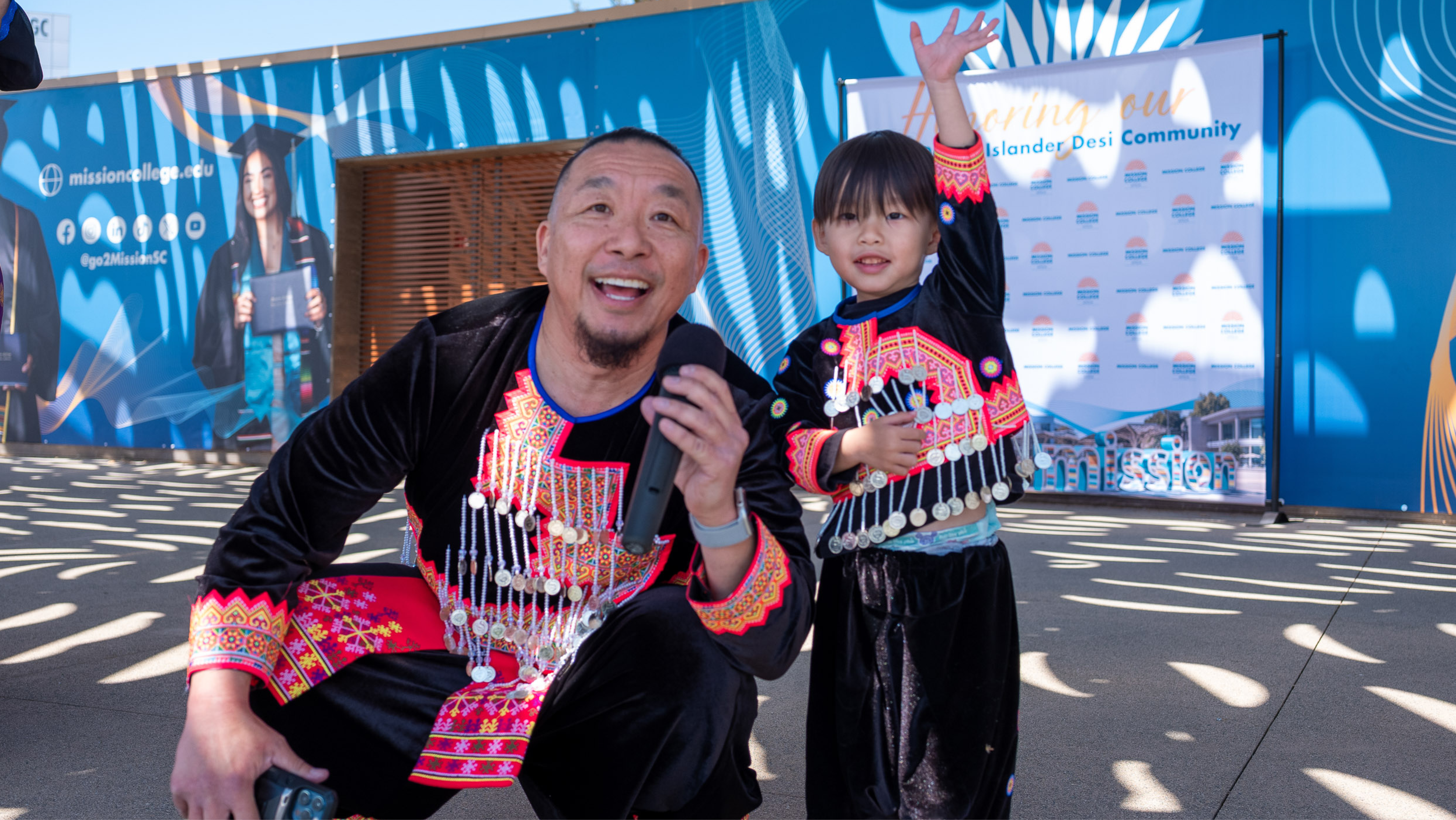 A smiling man crouching with a microphone alongside a toddler who is raising their hand; both are wearing matching traditional Hmong outfits featuring colorful embroidered trim and dangling silver coins.