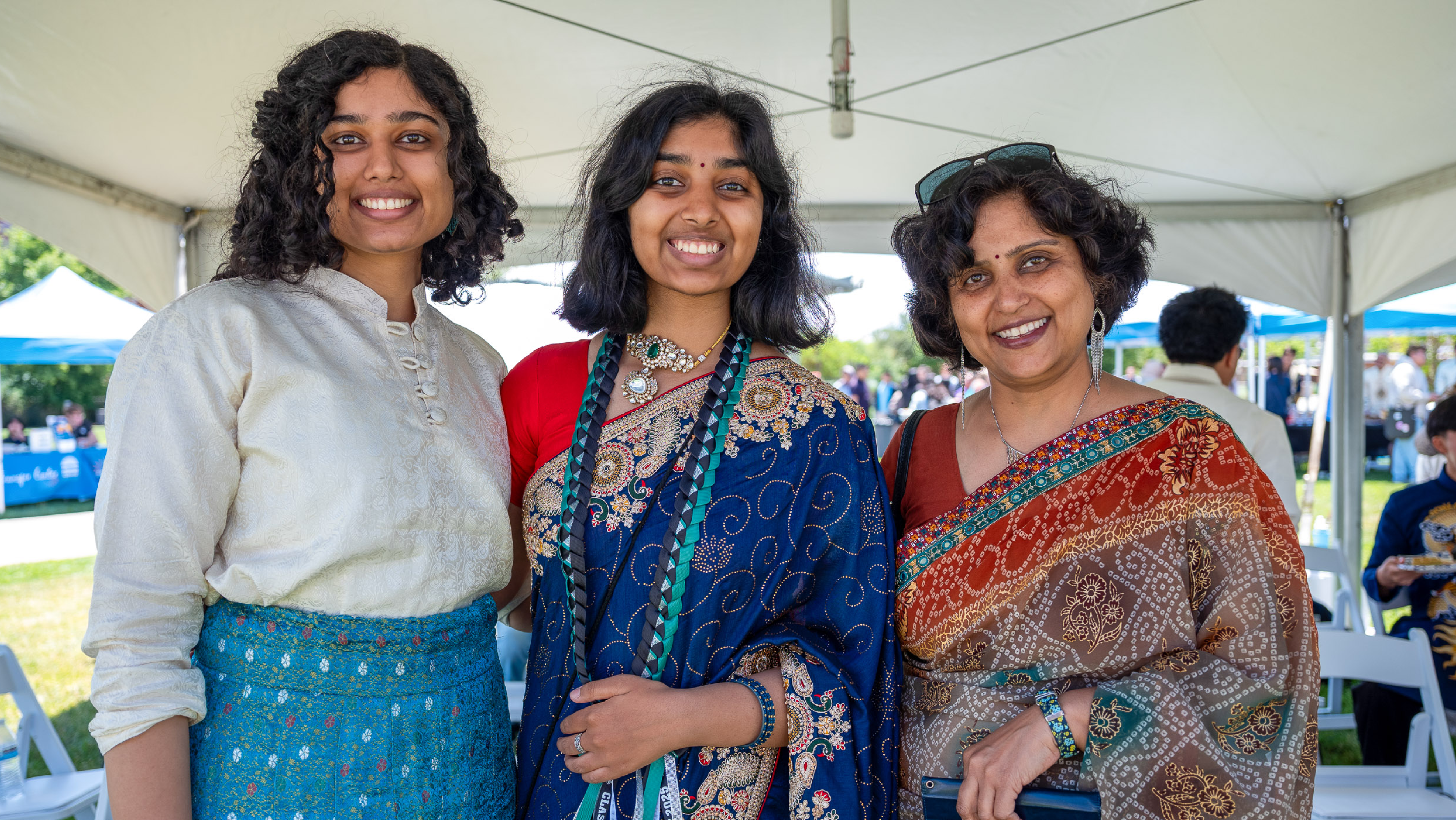 Three women smiling together under a white tent; the woman in the center wears an ornate royal blue saree with graduation sashes, flanked by two companions in traditional attire.