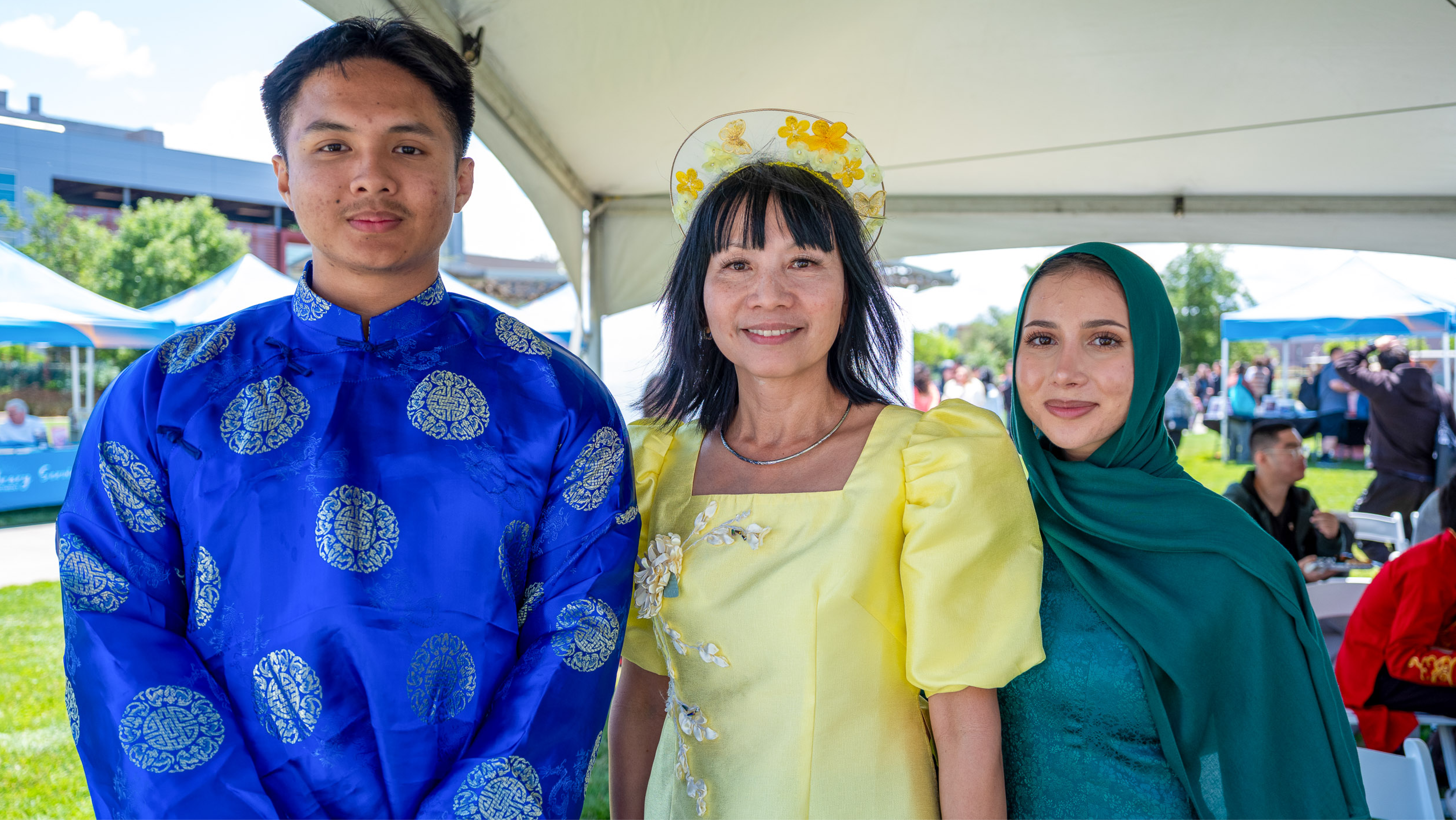 Three individuals posing outdoors: a young man in a bright blue patterned Áo Dài, a woman in a yellow dress with a floral headband, and a young woman wearing a teal top and matching green hijab.