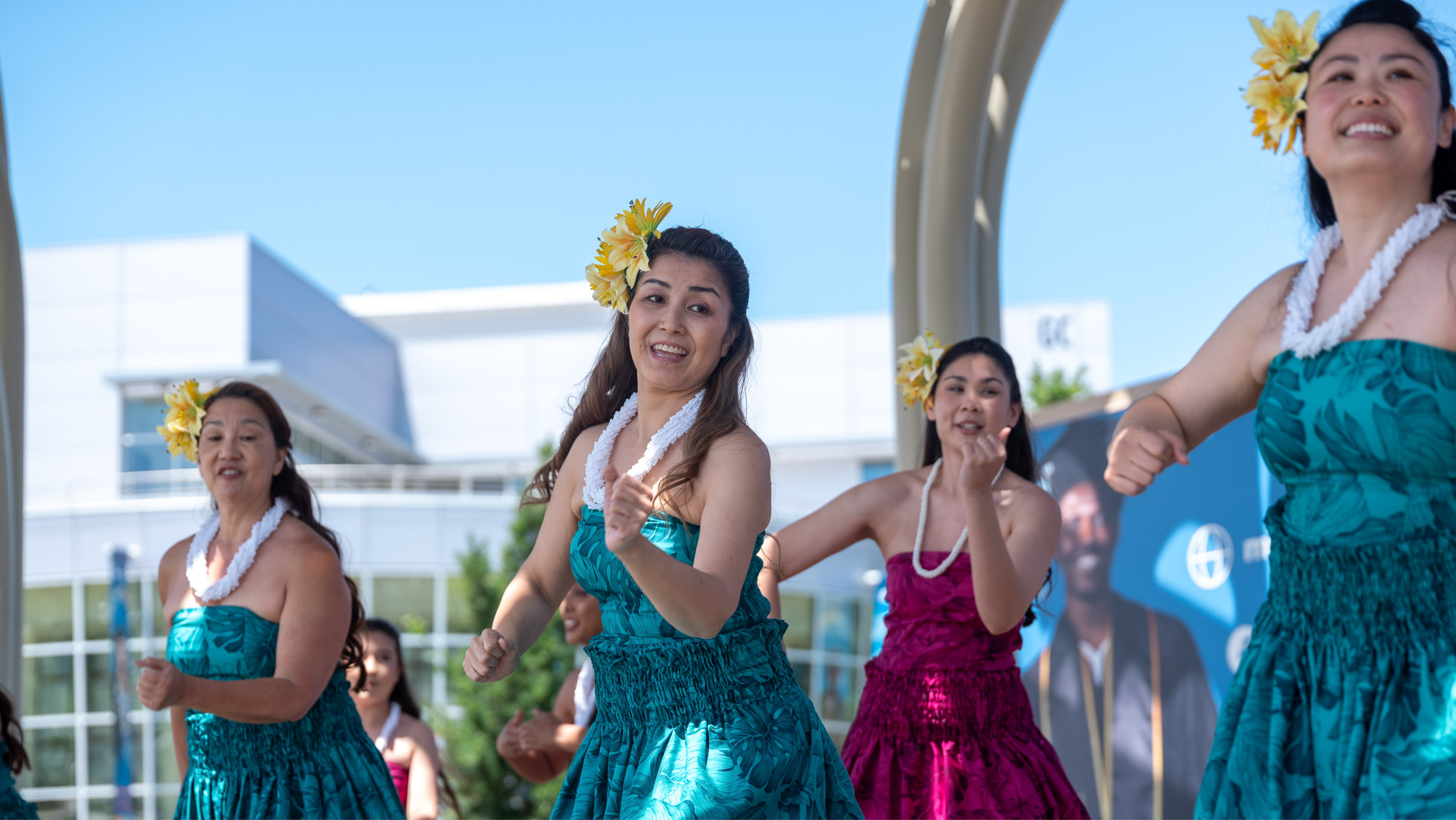 A group of dancers in matching teal floral strapless dresses, white lei necklaces, and yellow flowers in their hair performing a traditional Hula dance outdoors.