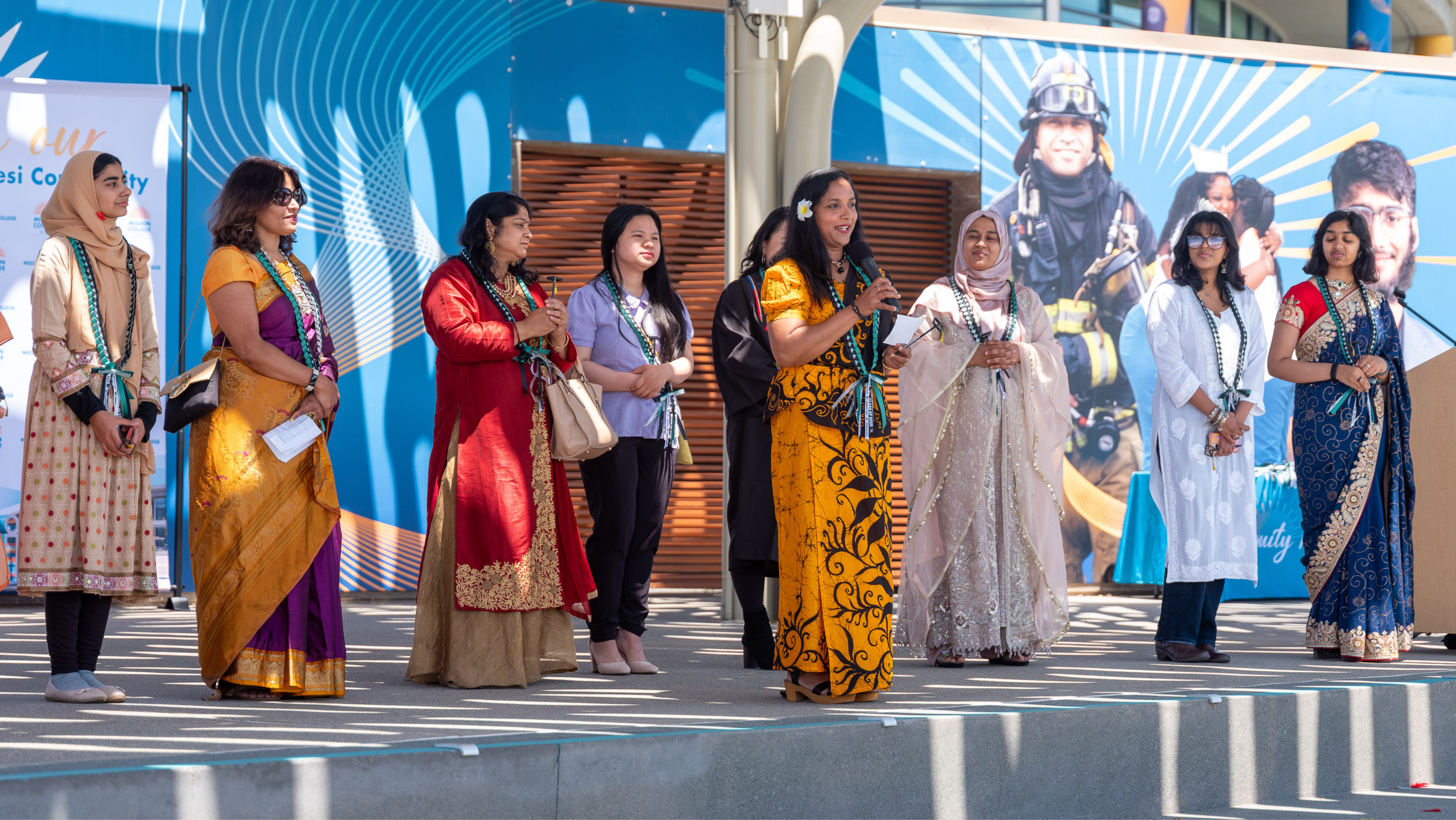 A line of women wearing vibrant sarees, salwar kameez, and traditional dresses standing on an outdoor stage, while one woman in a yellow patterned dress speaks into a microphone.
