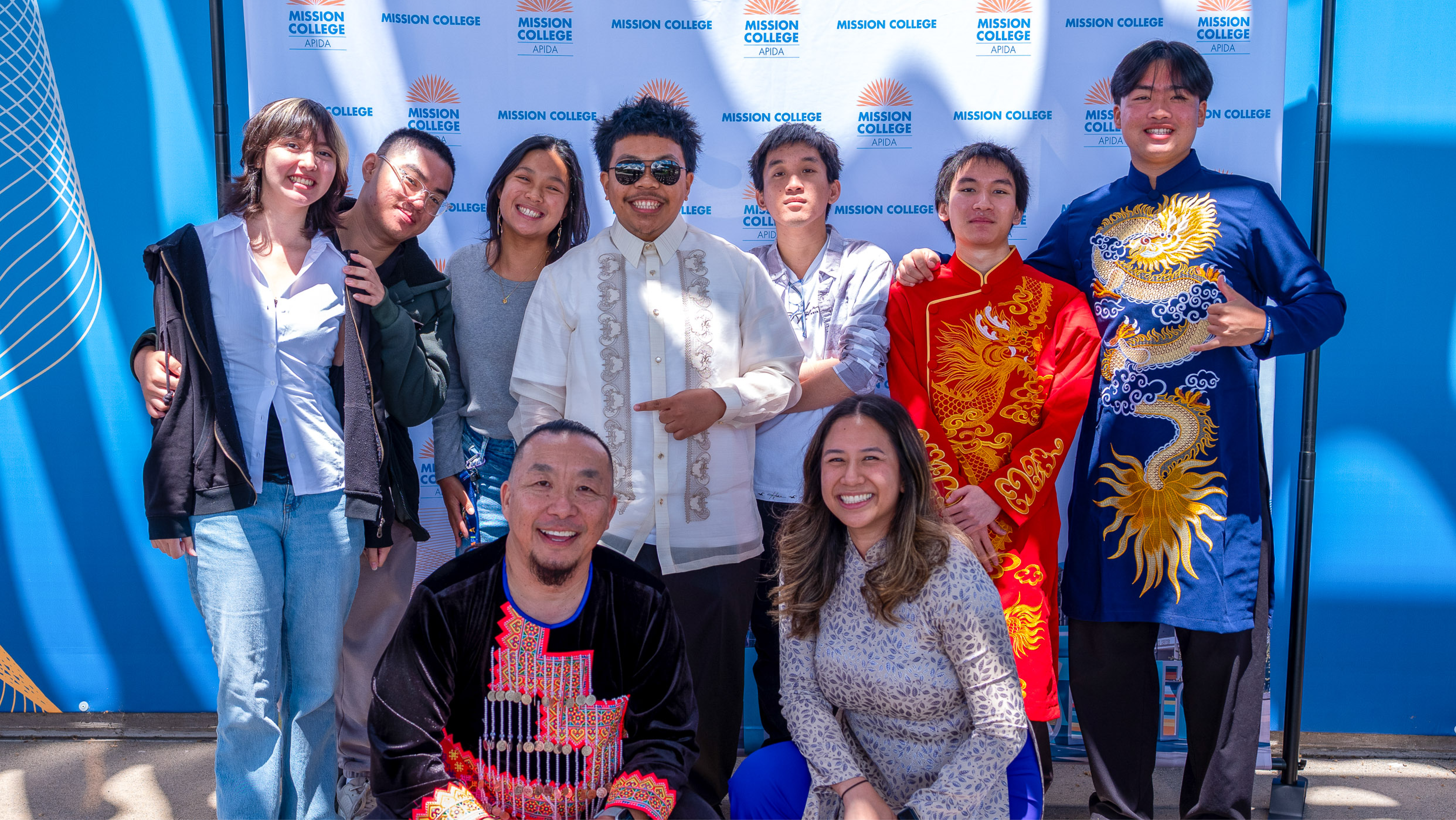 A group of people and presenters posing together in front of a blue Mission College APIDA backdrop, dressed in a mix of traditional cultural garments (including Barong Tagalog and Áo Dài) and casual clothing.