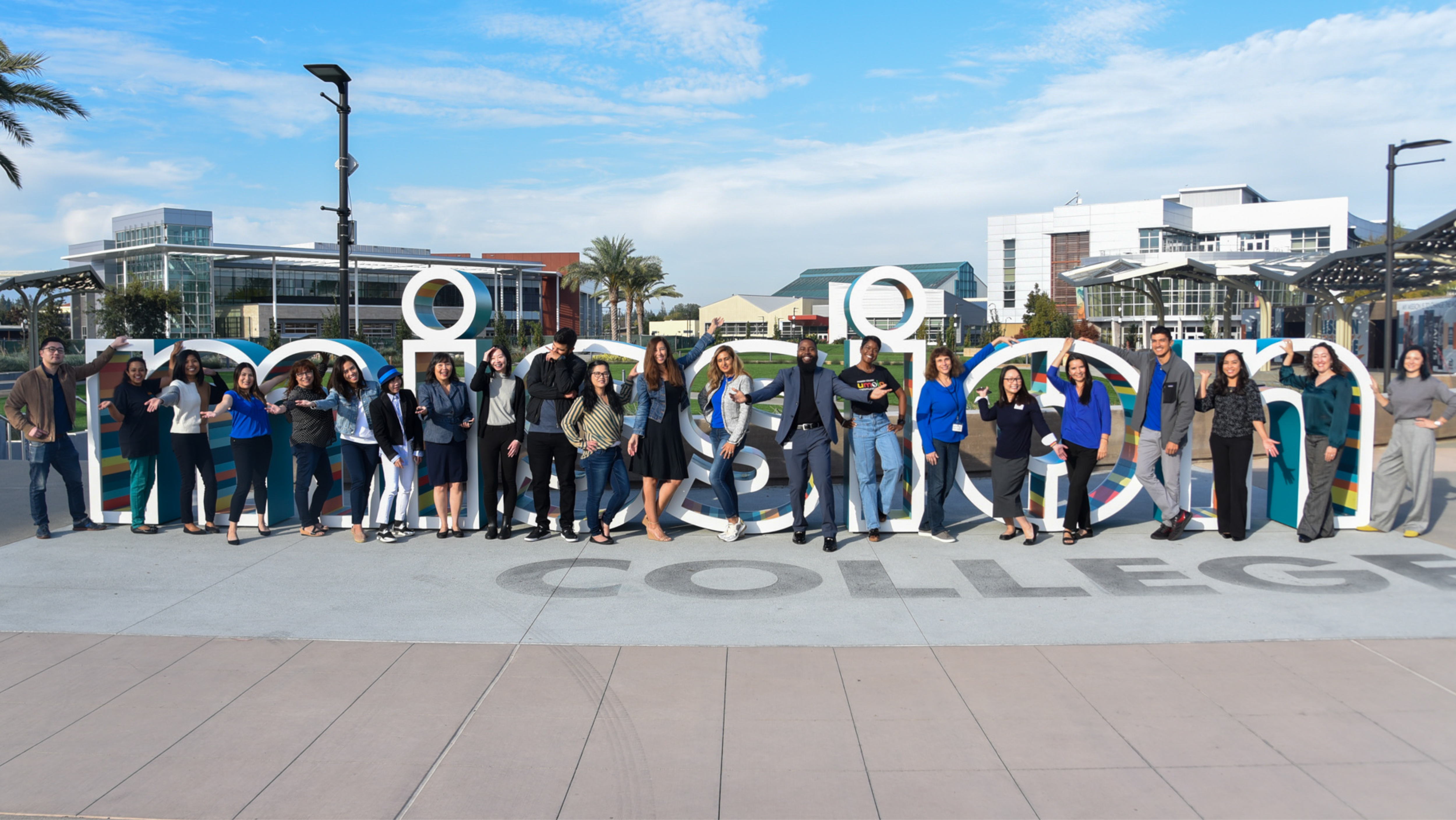A large group of Mission College students and staff posing enthusiastically around the large "MISSION" letter installation outdoors.