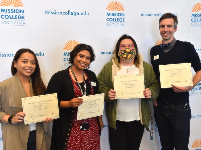 Vianey Topete holds an award/piece of paper with three colleagues in front of a backdrop decorated with the Mission College logo.