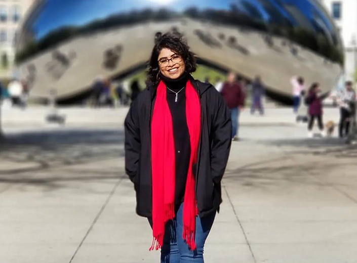 Vianey Topete, a woman of Latinx descent wears blue jeans, a black jacket, and a red scarf. She stands in front of a large metallic sphere/sculpture. She has short wavy black hair.