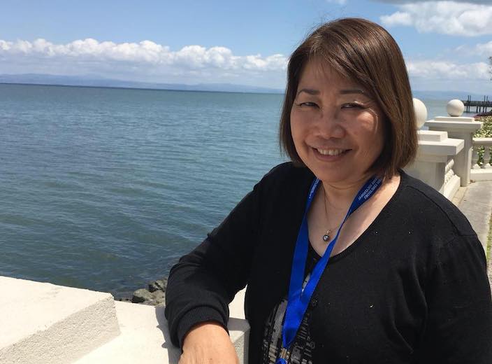 Ann Cowels, an Asian-American woman in her fifties, poses by a beautiful ocean on a white stone deck.