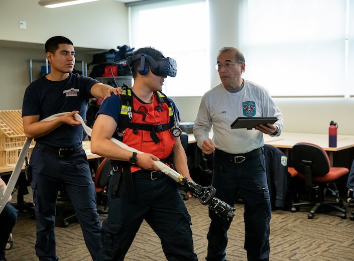 Two fire academy students practice a drill indoors. One wears a VR headset amd holds a long pole tool.