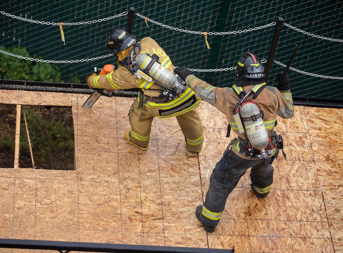 Two fire academy students practice a drill indoors. One wears a VR headset amd holds a long pole tool.