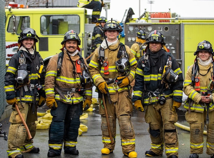 Group of firefighter cadets in gear at a drill. Four are men, one is a woman.