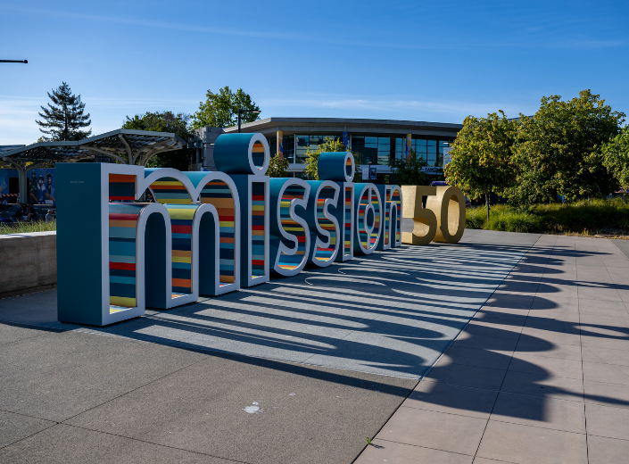 Large Mission College 50th anniversary sculpture in blue, yellow, and red decorative lettering sitting in a sunny campus plaza.